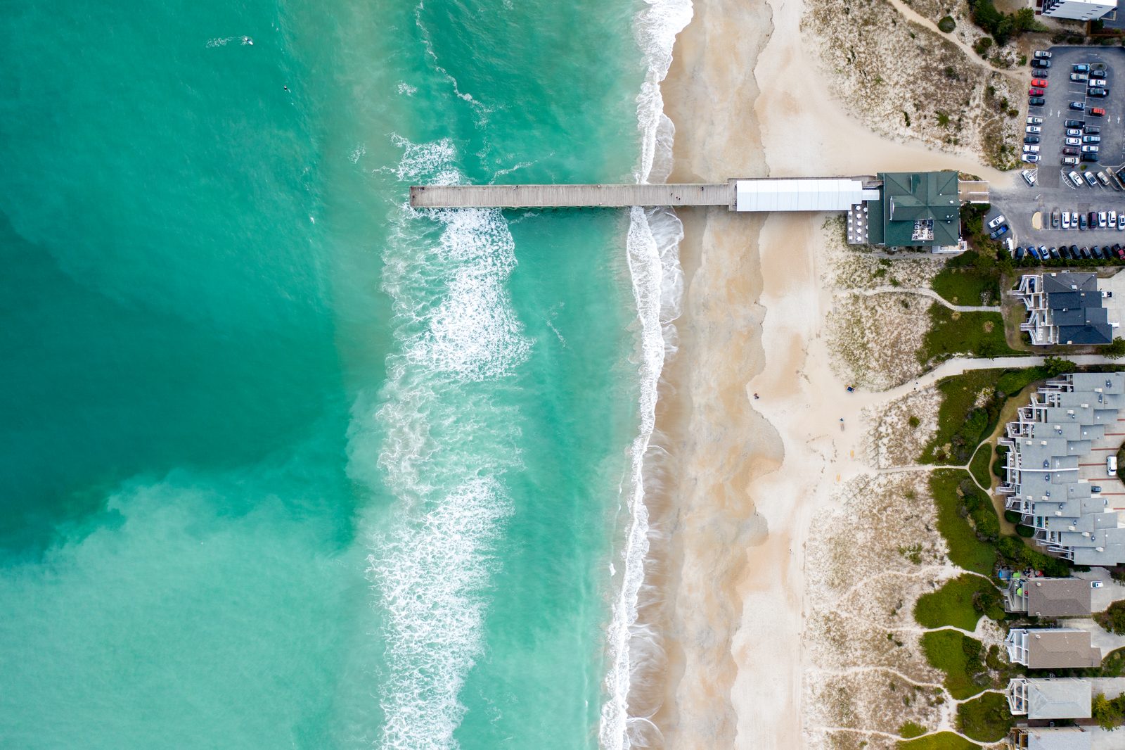 Aerial view of a coastal pier extending into turquoise ocean waves with beachfront homes and parking lot.
