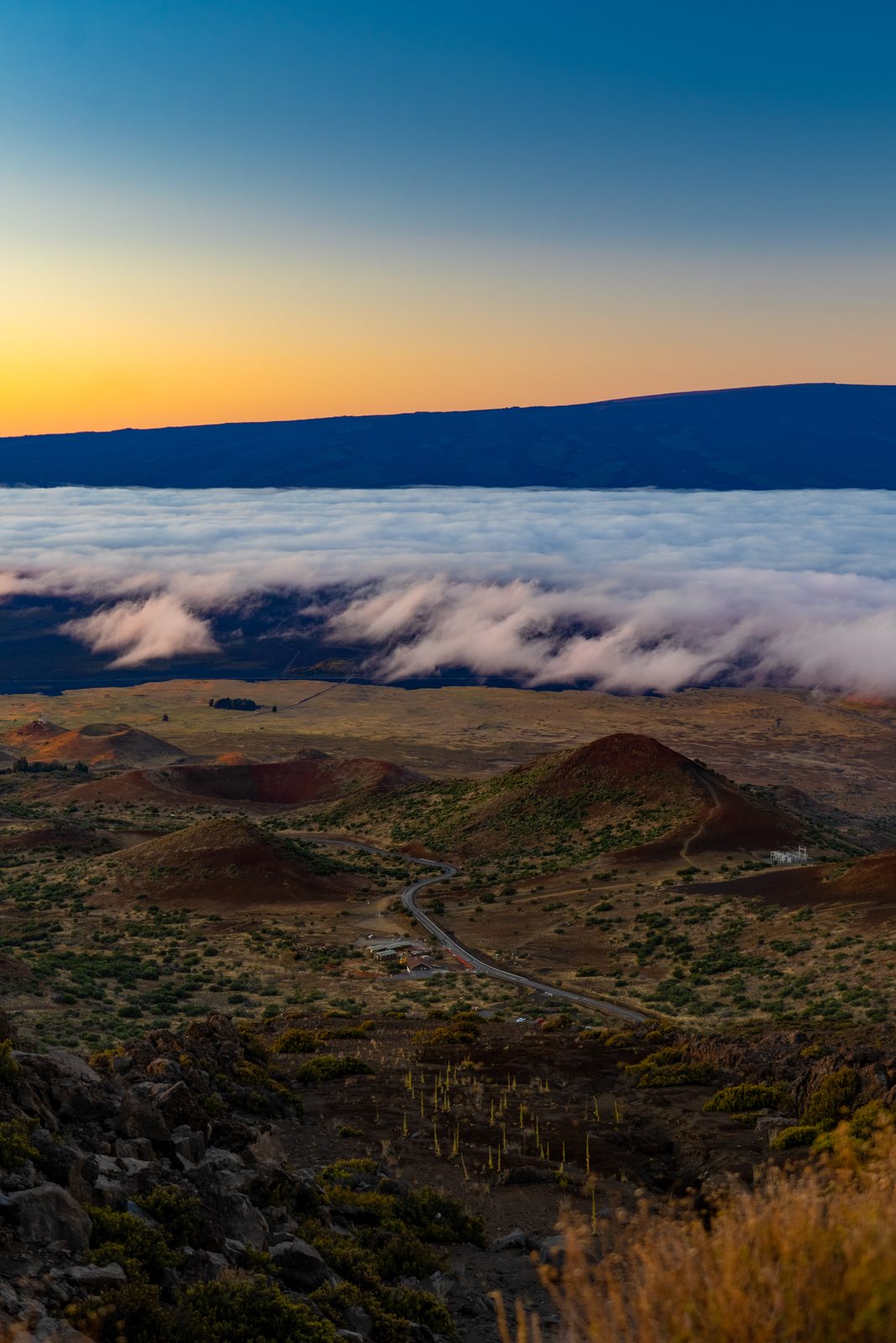 A winding road cuts through volcanic terrain beneath a sea of clouds at golden sunrise.