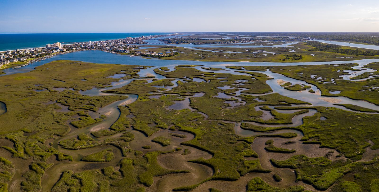 Aerial view of a sprawling salt marsh with winding tidal channels leading to a coastal town and ocean.
