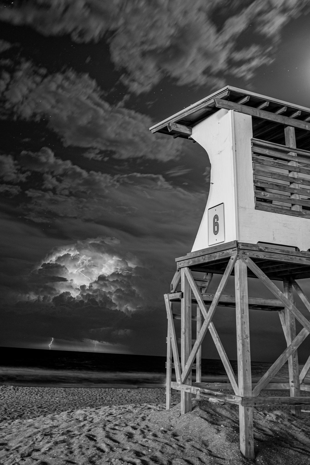 A lifeguard tower stands sentinel on an empty beach beneath a dramatic stormy sky with lightning.