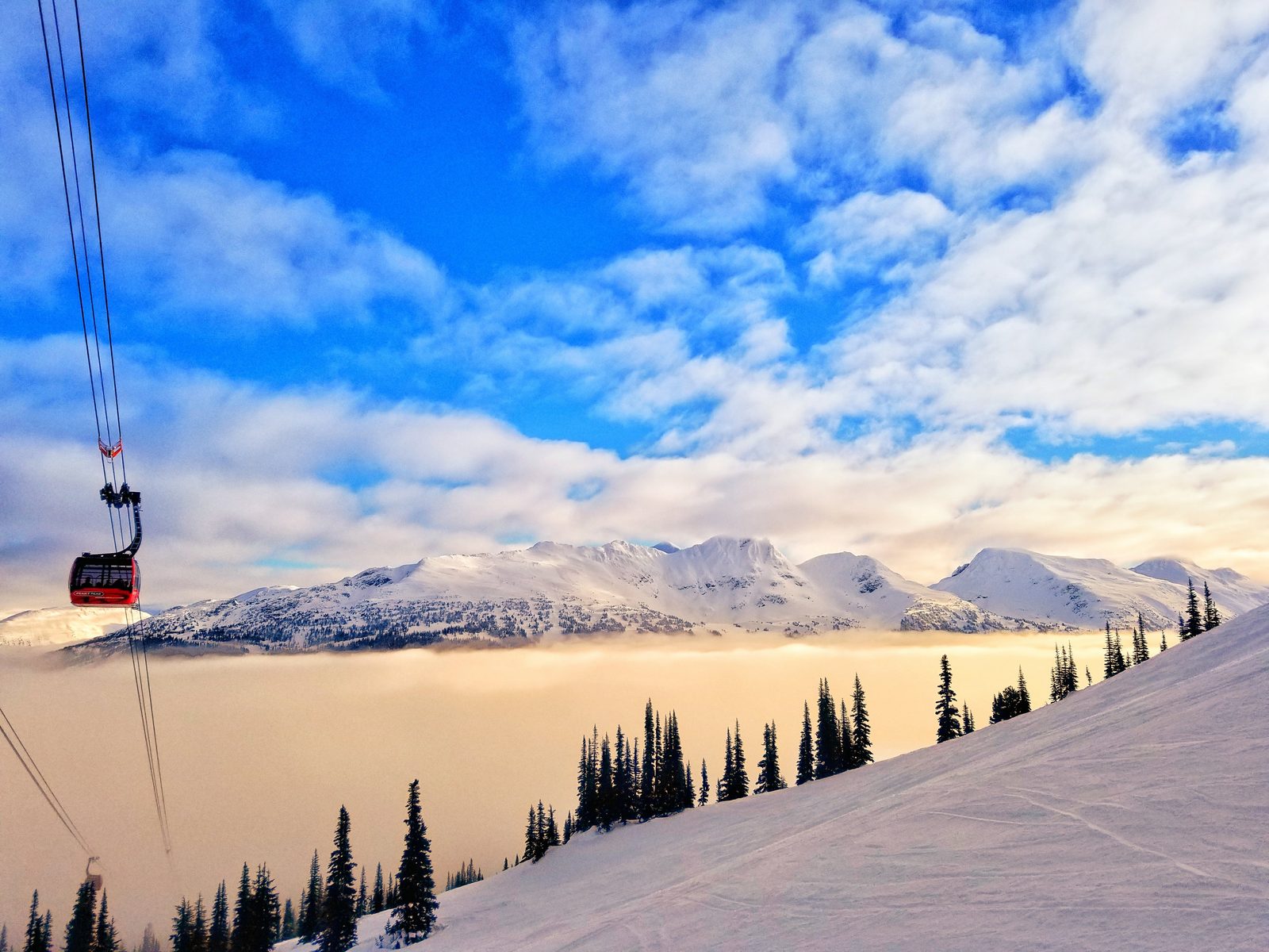 A red gondola cable car ascends a snowy mountain slope above a misty valley with evergreen forests.