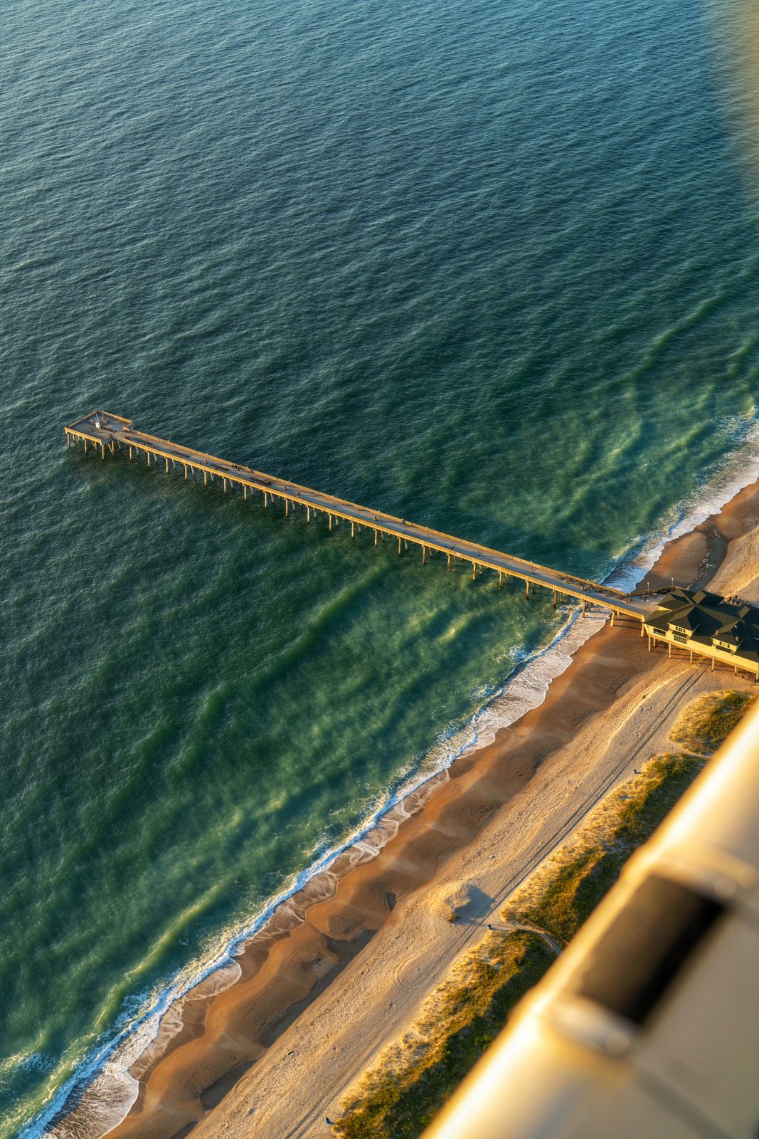 Aerial view of a weathered wooden pier extending into turquoise ocean waters beside a sandy beach.