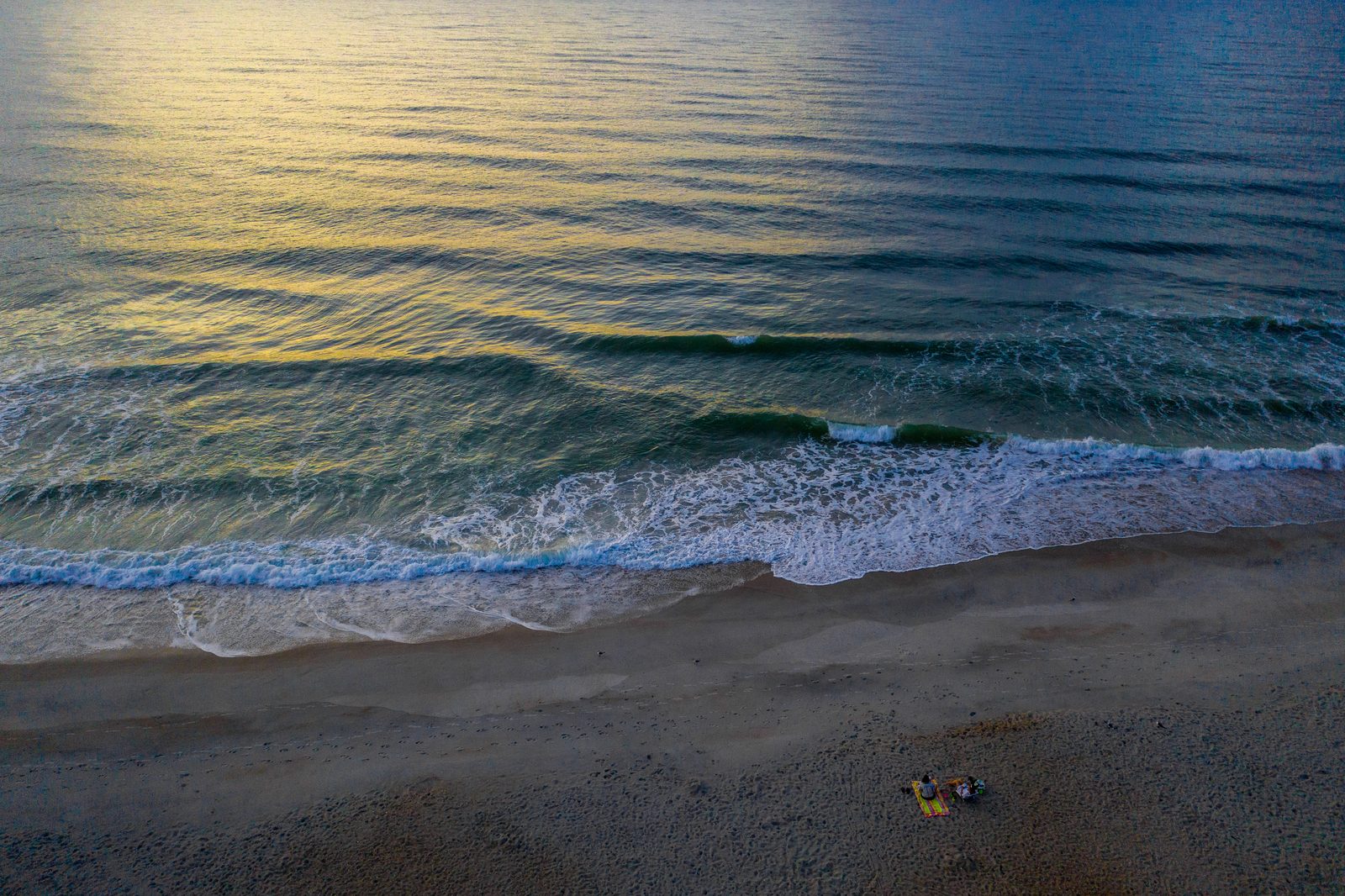 A solitary beachgoer relaxes on the sand as golden sunset light illuminates the rolling ocean waves.